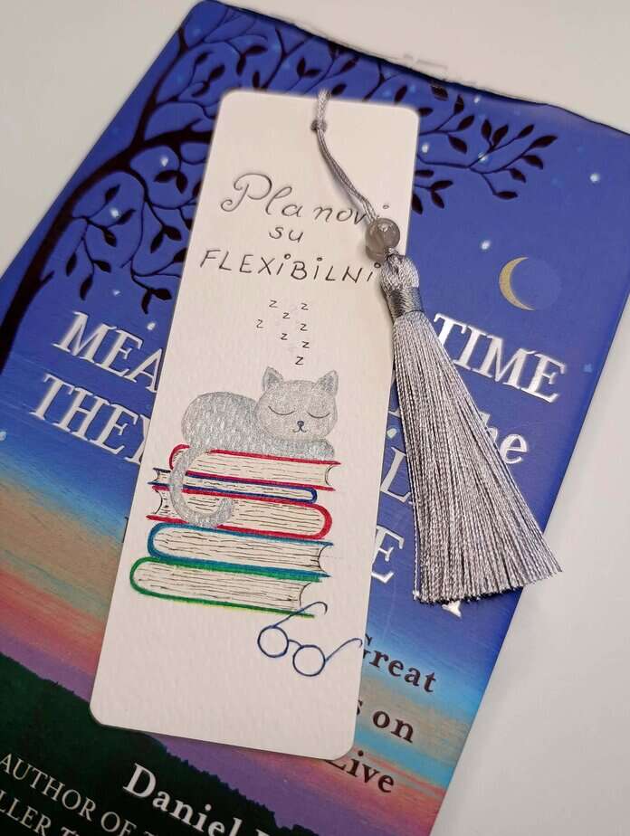 Ručno oslikani označivač stranica (bookmark) s motivom mačke koja spava na knjigama i tasselom, dimenzije 15x5, medij akvarel na 300 g/m² papiru. Idealno za ljubitelje knjiga.