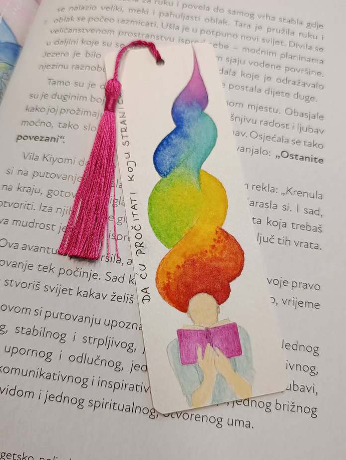 Ručno oslikani označivač stranica (bookmark) s motivom djevojke s kosom boje duge kako čita knjigu i tasselom, dimenzije 15x5, medij akvarel na 300 g/m² papiru. Idealno za ljubitelje knjiga.