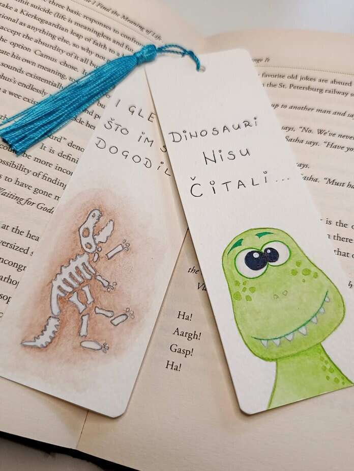 Ručno oslikani označivač stranica (bookmark) s motivom dinosaurom T-Rexom i tasselom, dimenzije 15x5, medij akvarel na 300 g/m² papiru. Idealno za ljubitelje knjiga.