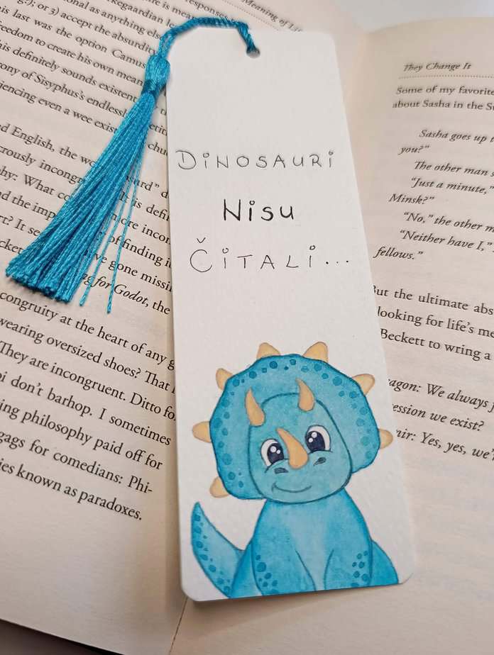 Ručno oslikani označivač stranica (bookmark) s motivom dinosaura Triceratopsom i tasselom, dimenzije 15x5, medij akvarel na 300 g/m² papiru. Idealno za ljubitelje knjiga.